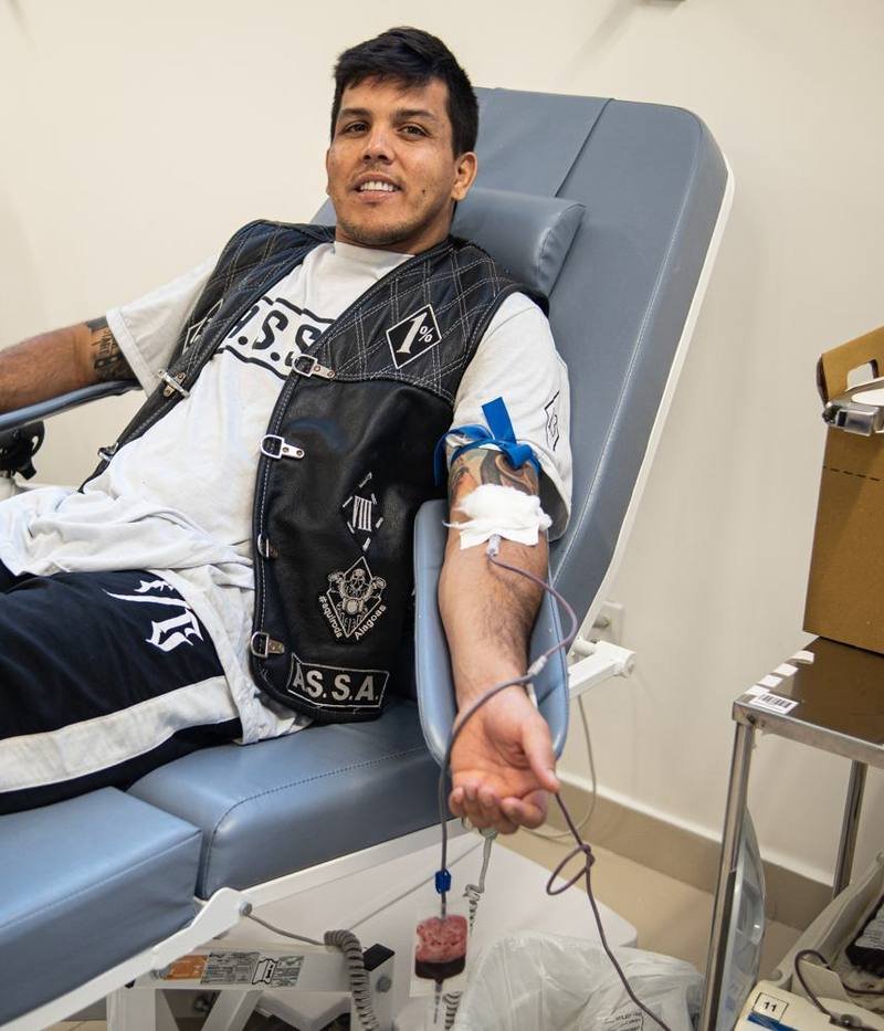 Motociclista doando sangue, com a bolsa enchendo ao lado da poltrona